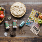 Apple pie with a basket of apples, vintage seed packets, and a magazine on a wooden surface.