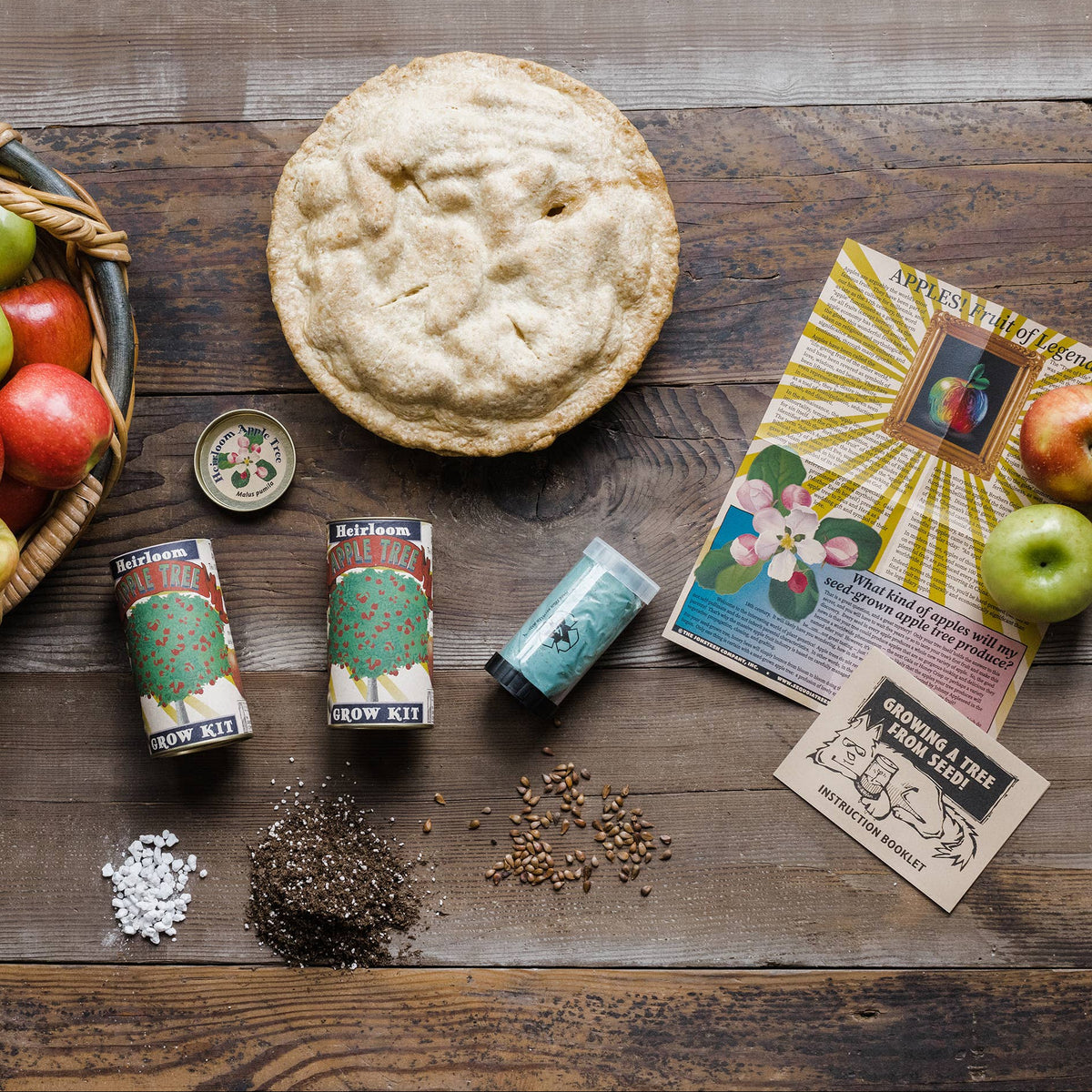Apple pie with a basket of apples, vintage seed packets, and a magazine on a wooden surface.