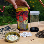 A hand holds a canister labeled Jurassic Tree next to a seed packet, perlite, soil, and a clear container on a wooden table with a garden background.