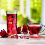 A glass of iced and a clear cup of hot Florida Cranberry - Hibiscus sabdariffa tea by Wellspring Gardens sit on a wooden table with fresh flowers, set against a blurred green outdoor background.