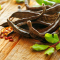 Wellspring Gardens Carob Bean (Ceratonia siliqua) pods and seeds, a natural cacao alternative, are displayed on rustic wood beside fresh green leaves.