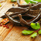 Wellspring Gardens Carob Bean (Ceratonia siliqua) pods and seeds, a natural cacao alternative, are displayed on rustic wood beside fresh green leaves.