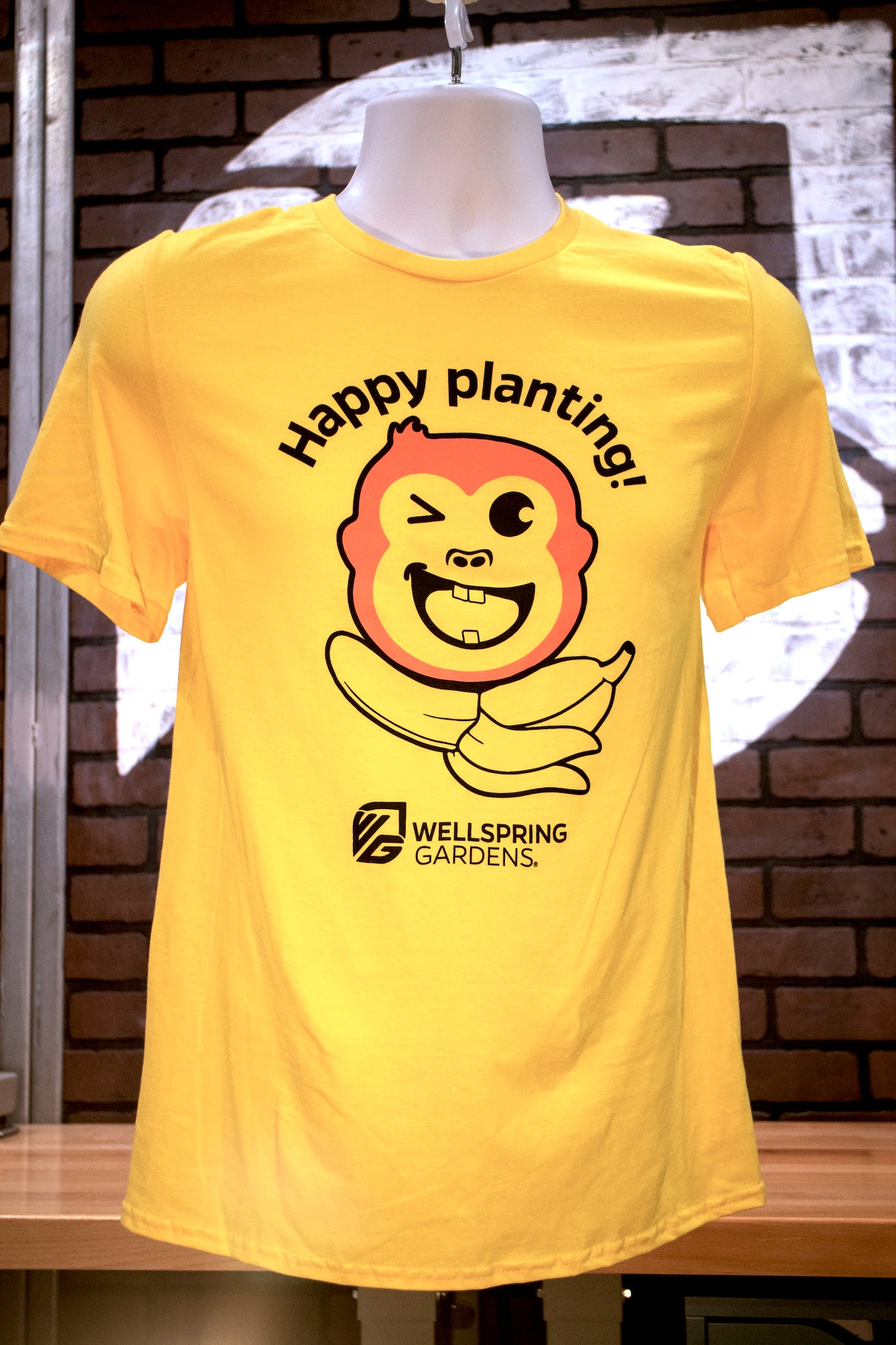 The Mekong Monkey 'Happy planting!' T-shirt by Wellspring Gardens features a winking monkey with a banana and “Happy planting!” text. The Wellspring Gardens logo appears below, making it ideal for fans of gardening mascots.