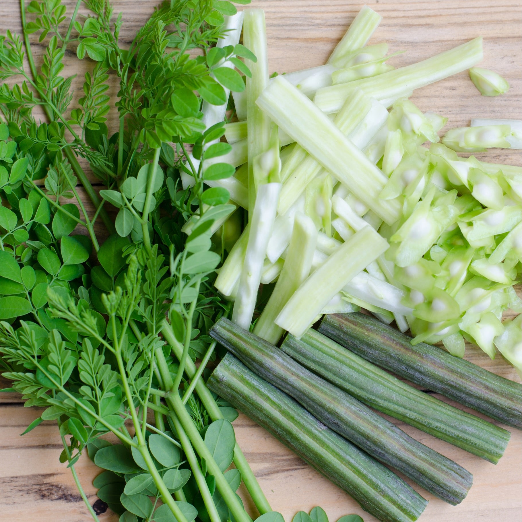 Wellspring Gardens' Moringa Tree (Ben Oil/Drumstick Tree) – Moringa oleifera displays fresh leaves, peeled stalks, and cut drumsticks on a light wooden surface, highlighting their vibrant green color and natural texture.