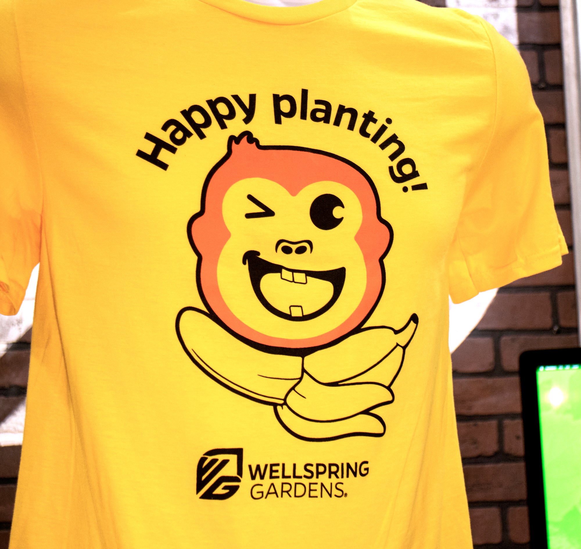 The Mekong Monkey 'Happy planting!' T-shirt by Wellspring Gardens is a yellow tee featuring the winking cartoon gardening mascot above a peeled banana tree, with "Happy planting!" and the Wellspring Gardens logo.
