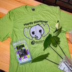The Wellspring Gardens Elepaio Elephant 'Happy planting!' T-shirt, in green, is displayed on a table next to plant leaves, fertilizer, and a plant cutting in a plastic bag.