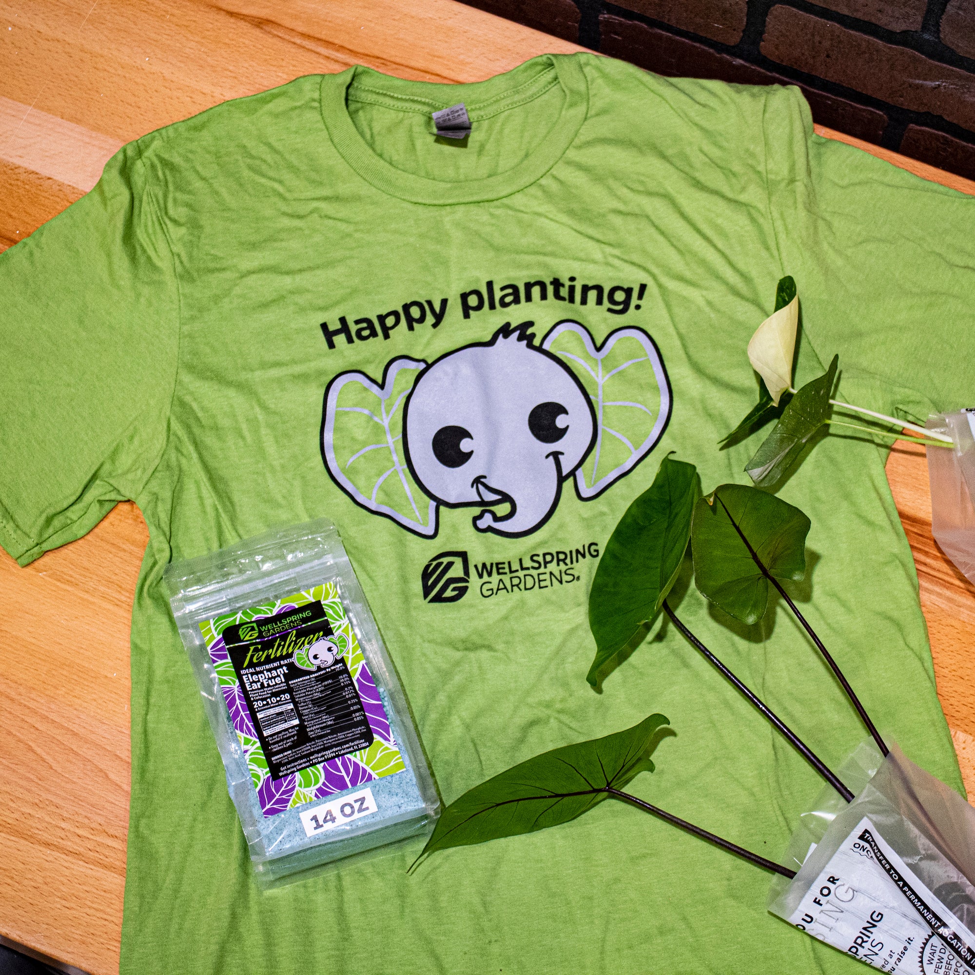 The Wellspring Gardens Elepaio Elephant 'Happy planting!' T-shirt, in green, is displayed on a table next to plant leaves, fertilizer, and a plant cutting in a plastic bag.