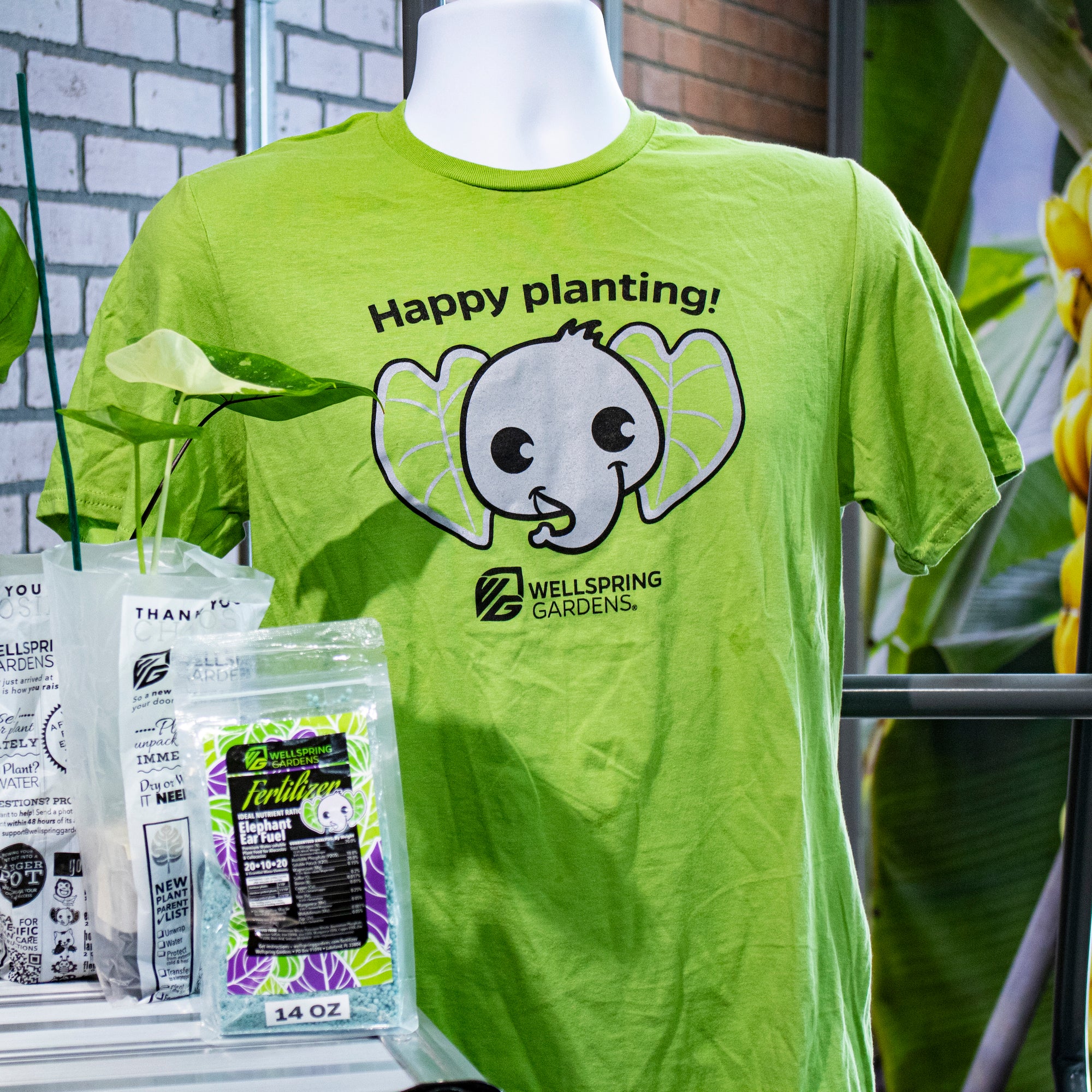 The Elepaio Elephant 'Happy planting!' T-shirt by Wellspring Gardens features an illustrated elephant face made of leaves and the phrase "Happy planting!"—the perfect gardening mascot among plant bags and lush greenery.